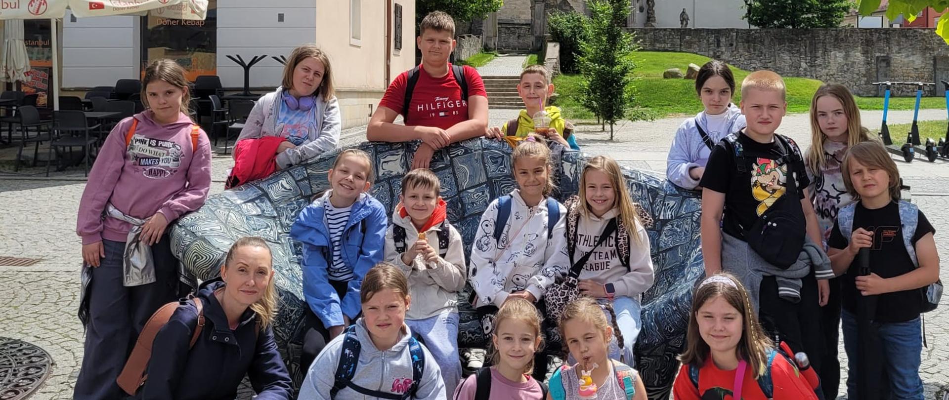 Grupowe zdjęcie uczestników warsztatów. Na fotografii znajdują się dzieci i młodzież, oraz jedna dorosła osoba. Uczestnicy pozują na zewnątrz, w kilku rzędach: część stoi, część siedzi, część kuca. Dzieci ubrane są swobodnie. W tle widoczny jest budynek, drzewa oraz trawa.