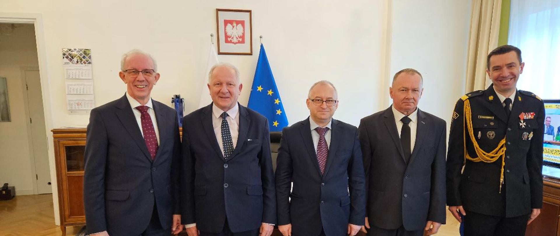 Pięciu mężczyzn elegancko ubranych, stoi obok siebie w gabinecie Podsekretarza Stanu MSWiA Wiesława Leśniakiewicza, uśmiechają się do zdjęcia. Na ścianie w tle wisi godło Polski oraz widoczna jest flaga Unii Europejskiej.