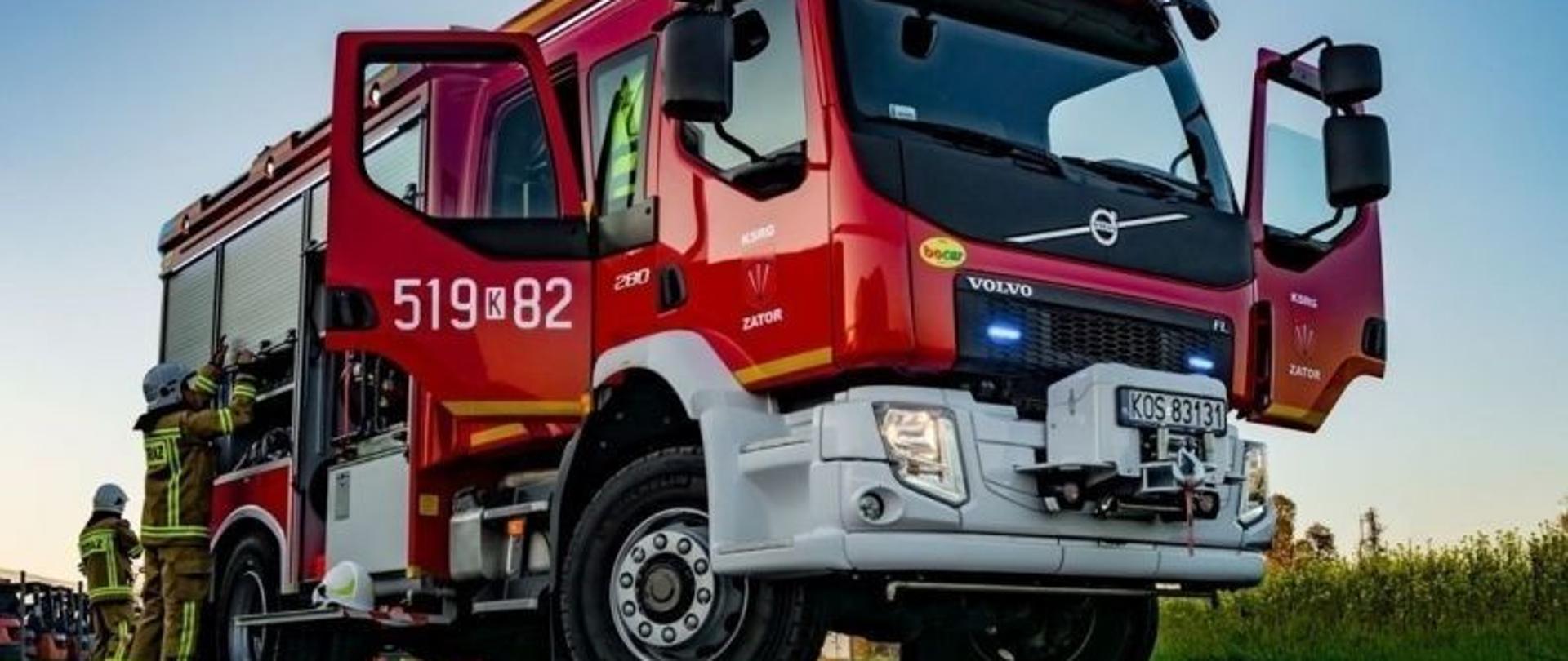 SAMOCHÓD OSP VOLVO zaparkowany na drodze asfaltowej, z otwartymi drzwiami od stroju kierowcy oraz tylne od strony pasażera. Z boku samochodu widać strażaków którzy otwierają boczną część schowka. 