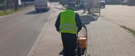 Pan w kamizelce odblaskowej zbiera liście z chodnika pchając pomarańczowy sprzęt odpowiedzialny za to