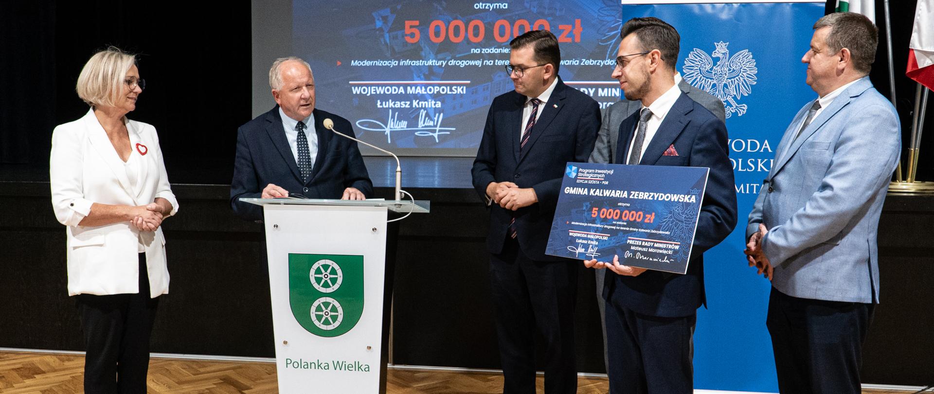 Osoby uczestniczące w przekazaniu symbolicznego czeku stoją wokół burmistrza Kalwarii, który przemawia przy białej mównicy. Za nimi utrzymane w granatowej kolorystyce banery i grafiki.