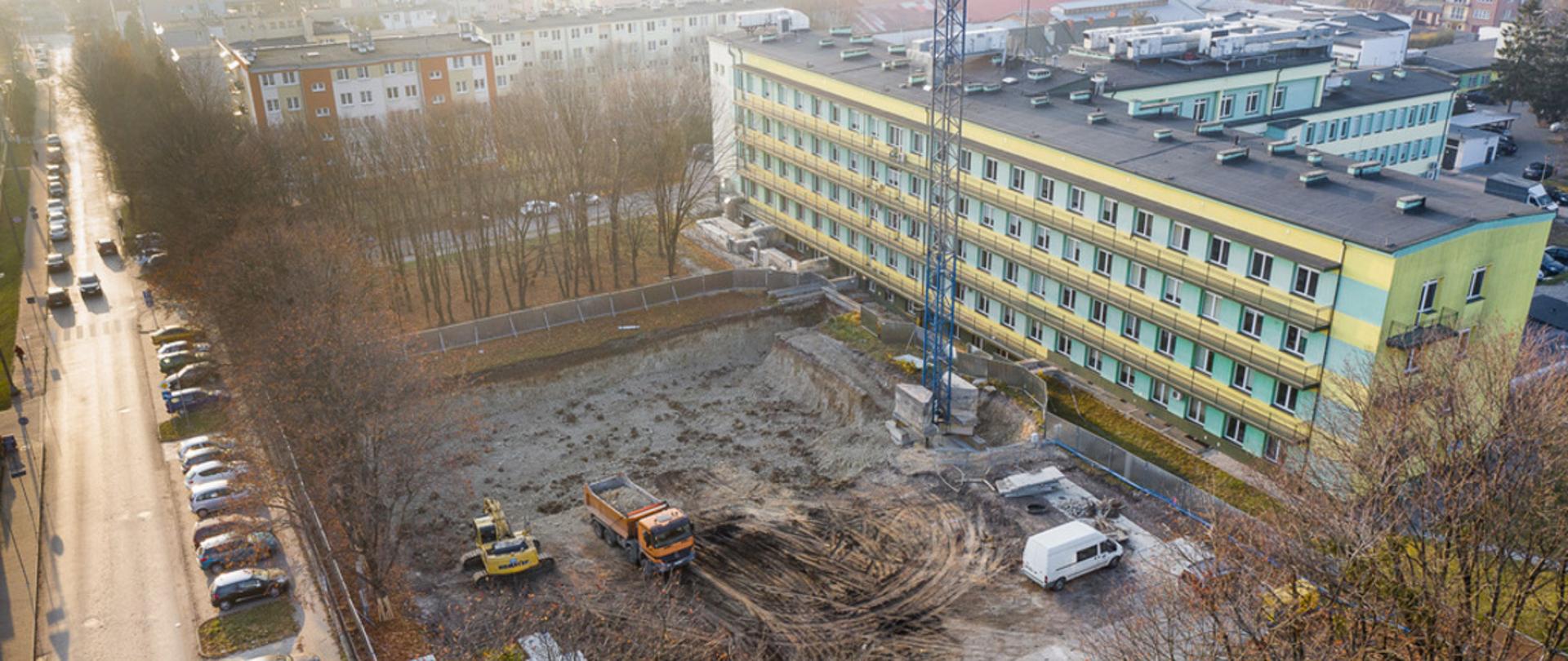 Zdjęcie przedstawia widok z lotu ptaka Samodzielnego Publicznego Zakładu Opieki Zdrowotnej w Kraśniku, a także teren budowy, gdzie wkrótce będzie dobudowany pawilon do szpitala. Na placu budowy jest dźwig, dwa samochody ciężarowe oraz bus.Teren jest rozkopany, przygotowany pod wylanie fundamentów. 