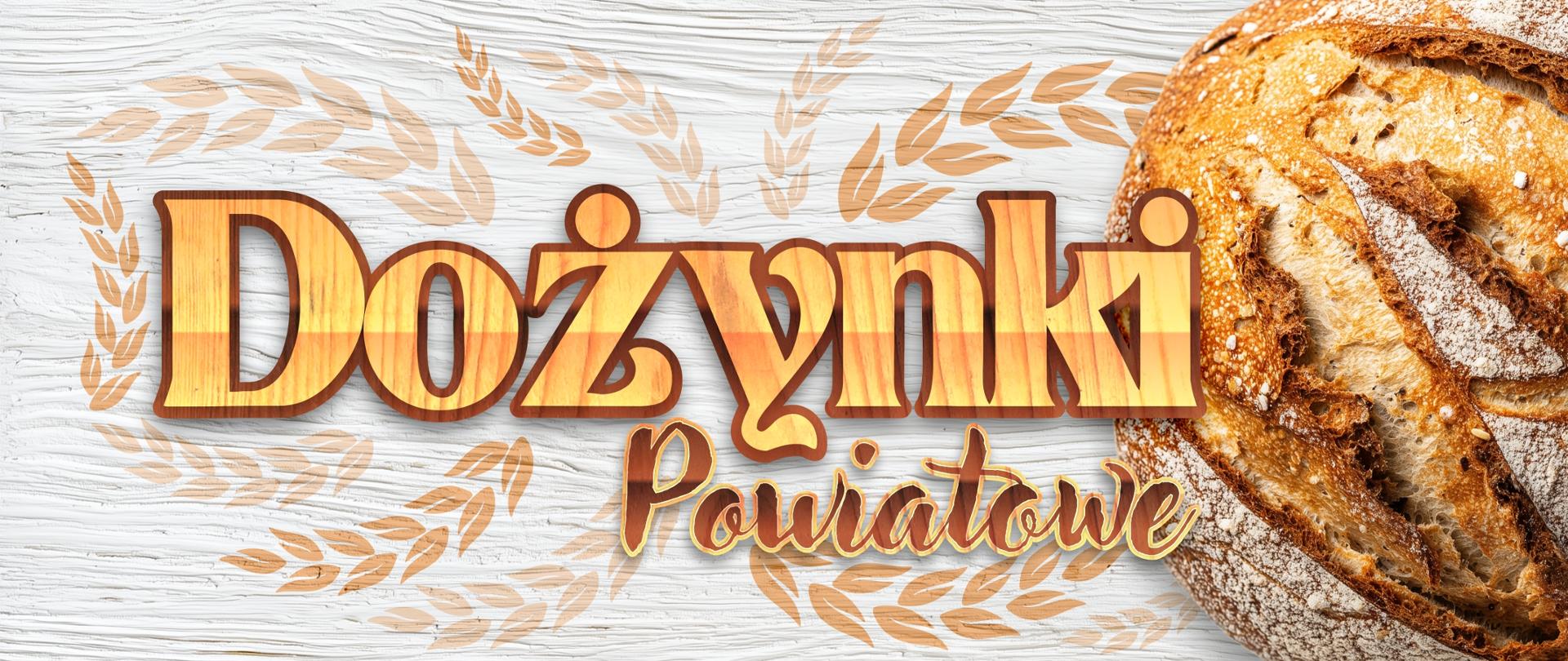 Grafika przedstawia ozdobny napis "Dożynki Powiatowe" otoczony wizerunkami kłosów. W tle znajduje się bochen chleba, który został ułożony na pomalowanej na biało desce.