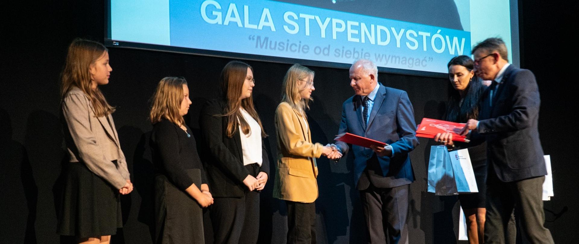Na czarnej podłodze sceny w sali widowiskowej stoją cztery uczennice i odbierają od burmistrza czerwone teczki, burmistrz ściska dłoń jednej z nich, po prawej prezes stowarzyszenie podaje teczki, za nimi ekran z napisem Gala Stypendystów Musicie od siebie wymagać.