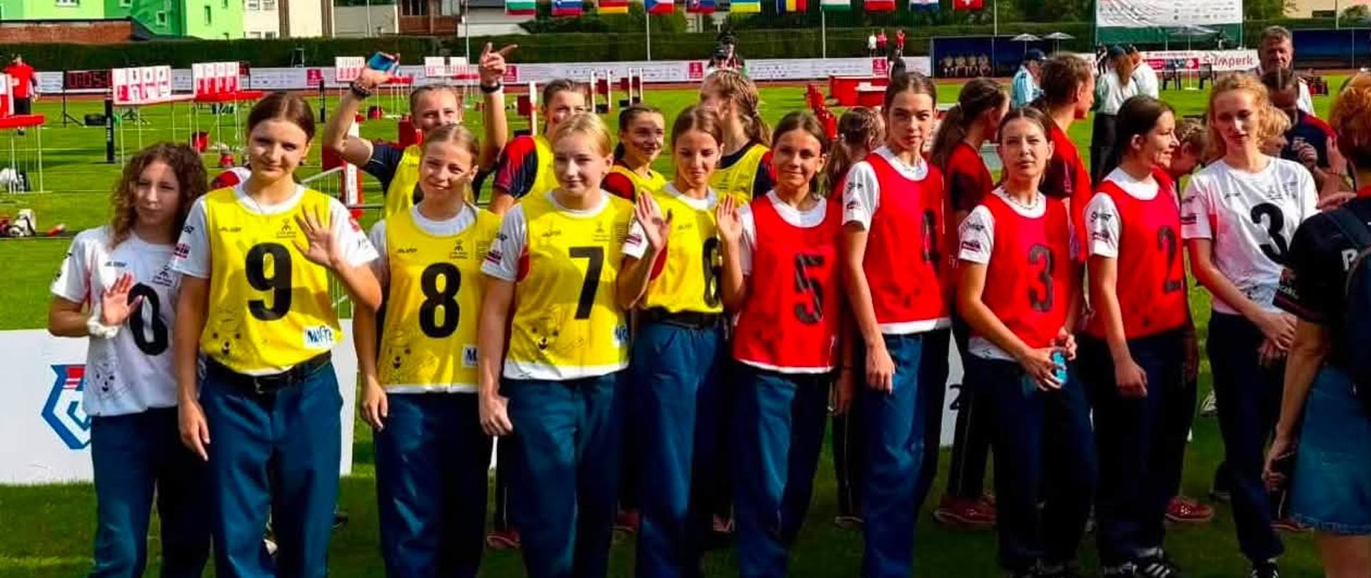 Grupa dziewcząt z młodzieżowych drużyn pożarniczych stojąca na stadionie podczas międzynarodowych zawodów CTIF w czeskim Šumperku. Uczestniczki mają na sobie stroje sportowe oraz kolorowe kamizelki z numerami, a w tle widoczne są flagi państw biorących udział w zawodach.