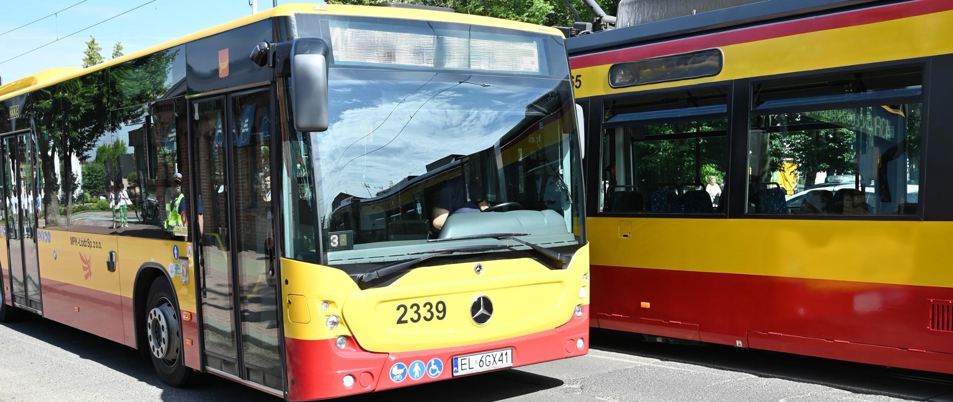 Żółto-czerwony autobus komunikacji miejskiej jedzie obok tramwaju w tych samych barwach. Oba pojazdy znajdują się na ulicy z torowiskiem tramwajowym, w słoneczny dzień. W tle widać drzewa, sieć trakcyjną oraz chodnik biegnący wzdłuż jezdni. Zdjęcie przedstawia autobusową komunikację zastępczą kursującą podczas wstrzymania ruchu tramwajów.