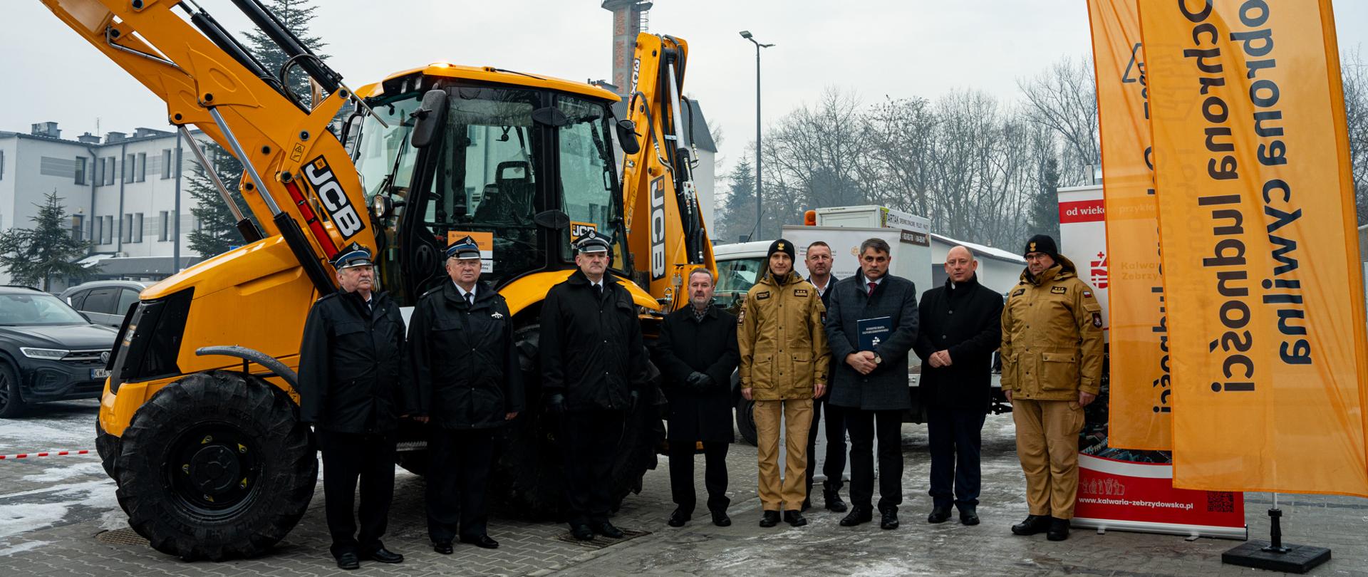 Grupowe zdjęcie uczestników uroczystego przekazania sprzętu w Kalwarii Zebrzydowskiej. Przedstawiciele administracji rządowej, samorządowej oraz służb pożarniczych stoją na placu przed nową koparko-ładowarką koloru żółtego. Obok widoczne są flagi programu ochrony ludności i obrony cywilnej. W tle zabudowania miejskie, zaparkowane pojazdy oraz zimowa aura.
