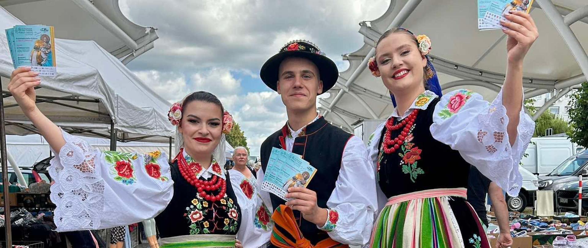 Dwie kobiety i mężczyzna w łowickich strojach ludowych z ulotkami w dłoniach na targowisku miejskim.