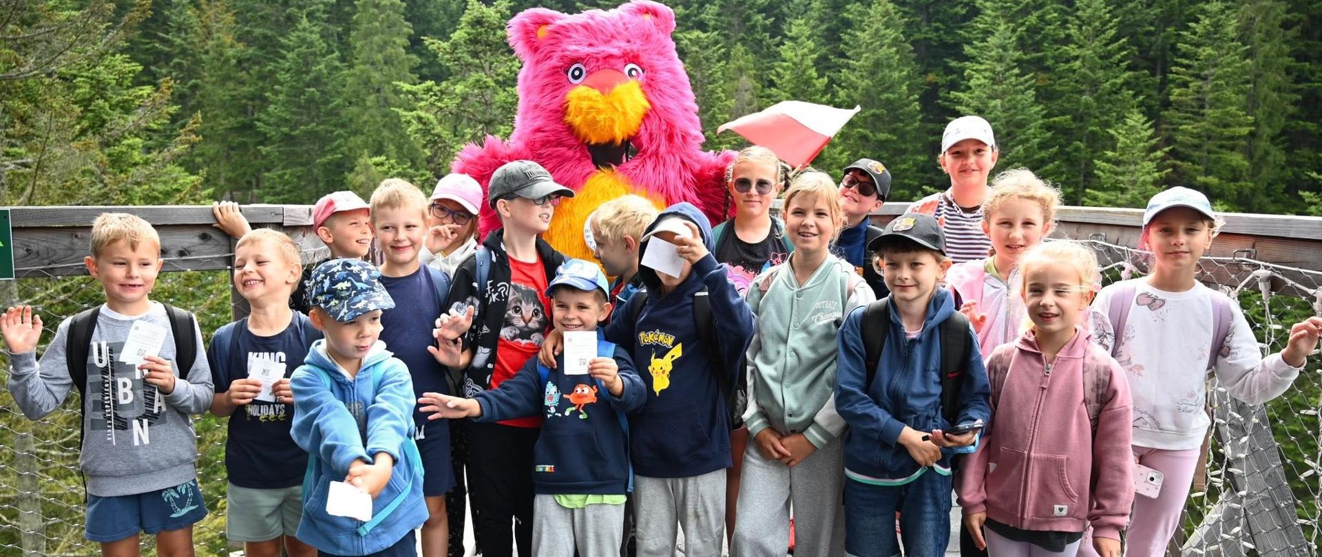 Grupa dzieci uczestniczących w wakacyjnym wyjeździe organizowanym przez Centrum Kultury, Sportu i Turystyki w Kalwarii Zebrzydowskiej pozuje do wspólnego zdjęcia na platformie widokowej wśród gór i lasów. Za dziećmi stoi różowa maskotka, a w tle widoczne są drzewa oraz polska flaga.