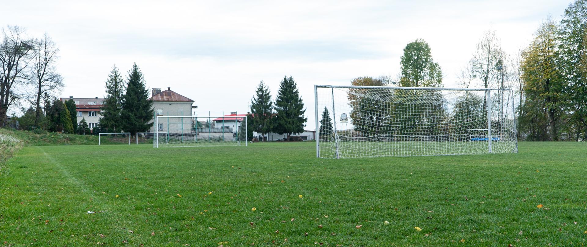 Na zielonej murawie boiska w centrum zdjęcia stoją dwie bramki służące do gry w piłkę nożną w tle znajduję się strefa kibica,dwie bramki,drzewa oraz budynki