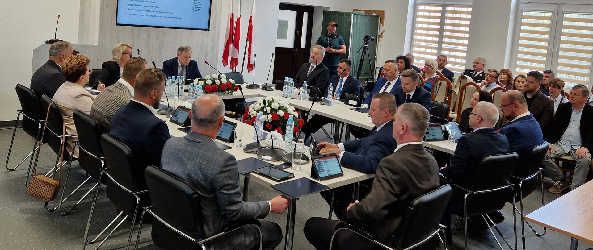 Wokół stołu zgromadzeni Radni Rady Miejskiej Kalwarii Zebrzydowskiej. w tle monitor z wynikami głosowania oraz trzy flagi państwowe.