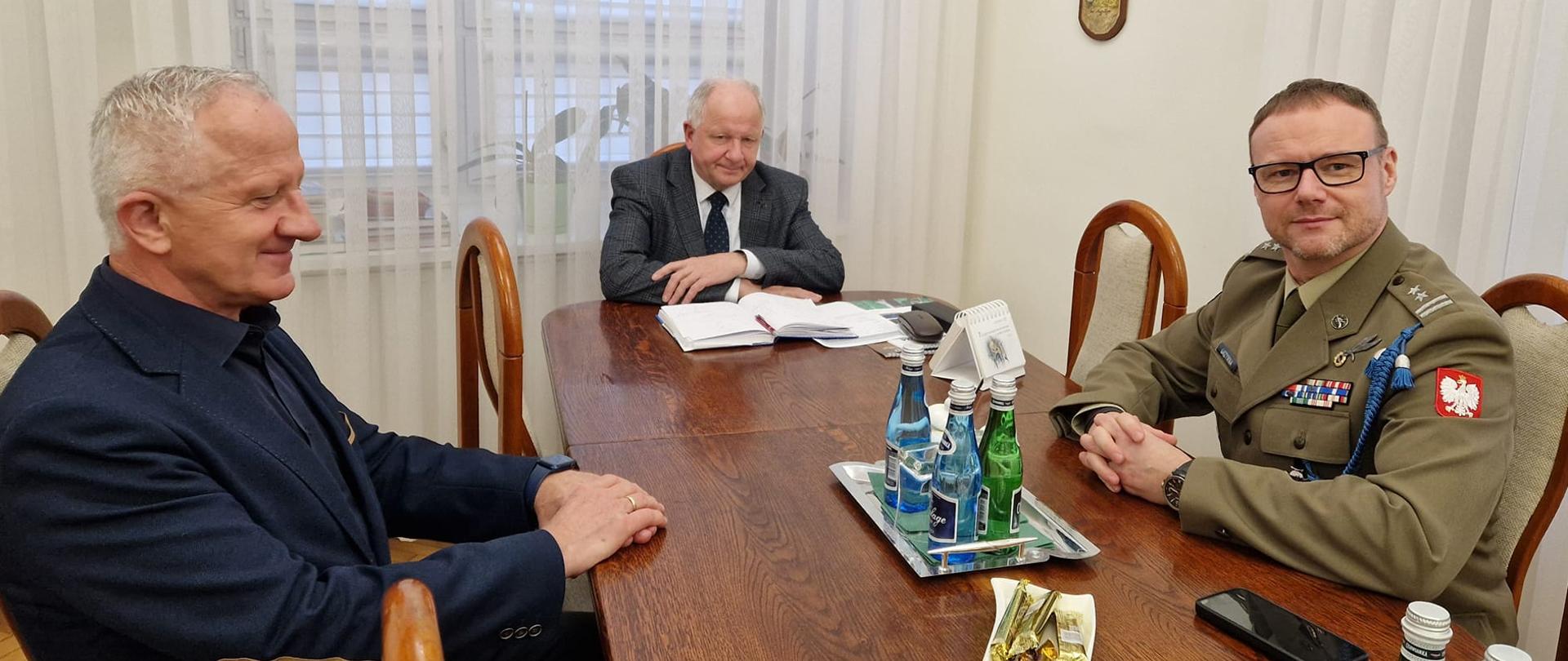 Spotkanie przy stole konferencyjnym trzech mężczyzn. Jeden mężczyzna to wojskowy, siedzi po prawej stronie. Na końcu stołu mężczyzna w garniturze i krawacie. Po lewej mężczyzna elegancki w Garniturze granatowym.