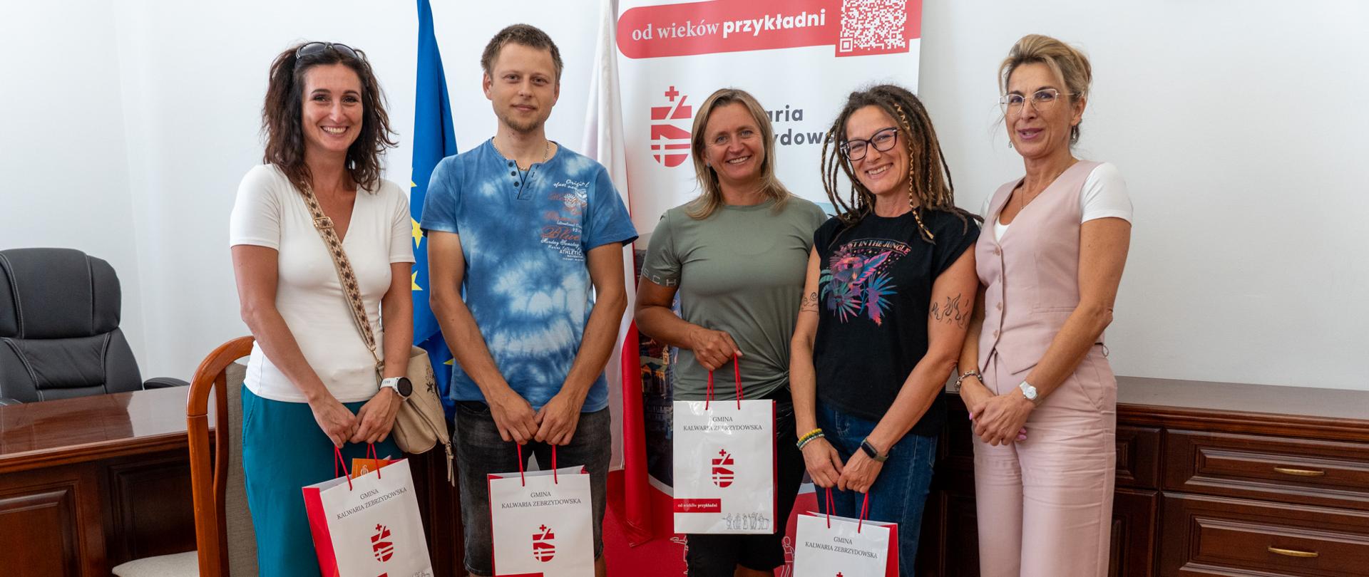 Zdjęcie grupowe laureatów rywalizacji „Kręcę dla Kalwarii” wraz z Sekretarz Gminy Kalwaria Zebrzydowska Renatą Borończyk podczas spotkania w Urzędzie Miasta. Uczestnicy trzymają torby z upominkami oznaczone herbem gminy. W tle widoczne są flagi oraz baner promocyjny Kalwarii Zebrzydowskiej.