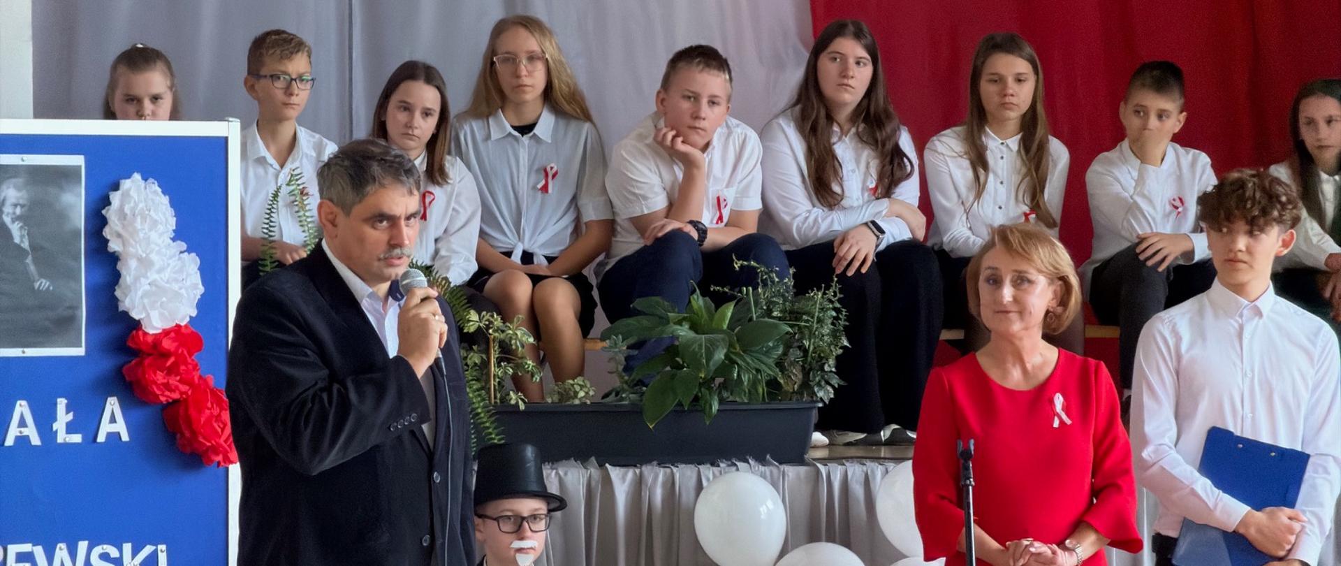 Na zdjęciu widać uroczystość szkolną z okazji święta narodowego. Na tle dużej biało-czerwonej dekoracji stoi mężczyzna przemawiający do mikrofonu, ubrany w ciemny garnitur. Obok niego stoi kobieta w eleganckiej czerwonej sukni. Za nimi siedzi grupa uczniów w odświętnych, biało-czarnych strojach, część z nich ma przypięte kokardki narodowe. Z boku widoczna jest tablica dekoracyjna z biało-czerwonymi kwiatami i wizerunkiem Stefana Żeromskiego. Atmosfera jest podniosła i nawiązuje do patriotycznego charakteru wydarzenia.