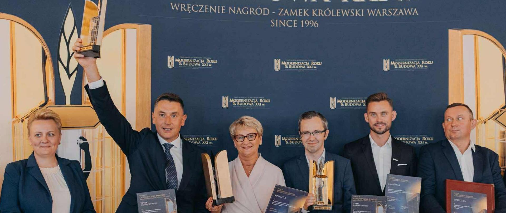 Grupa sześciu osób w eleganckich strojach stoi na tle ścianki z napisem „Modernizacja Roku & Budowa XXI w.”. Trzymają w rękach statuetki i dyplomy odebranych nagród. Jeden z mężczyzn unosi puchar w górę z uśmiechem.