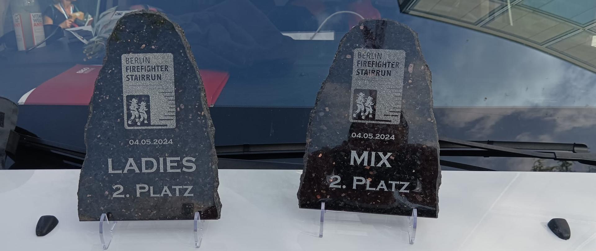 Na białej masce samochodu z czerwonym napisem straż stoją dwa kamienne puchary. Puchary z wyciętego granitu. na pucharze z lewej strony napis: Berlin Firefighter Stairrun 04.05.2024, poniżej Ladies 2. Platz. Na drugim identycznym pucharze napis: Berlin Firefighter Stairrun 04.05.2024, poniżej MIX 2 Platz.
