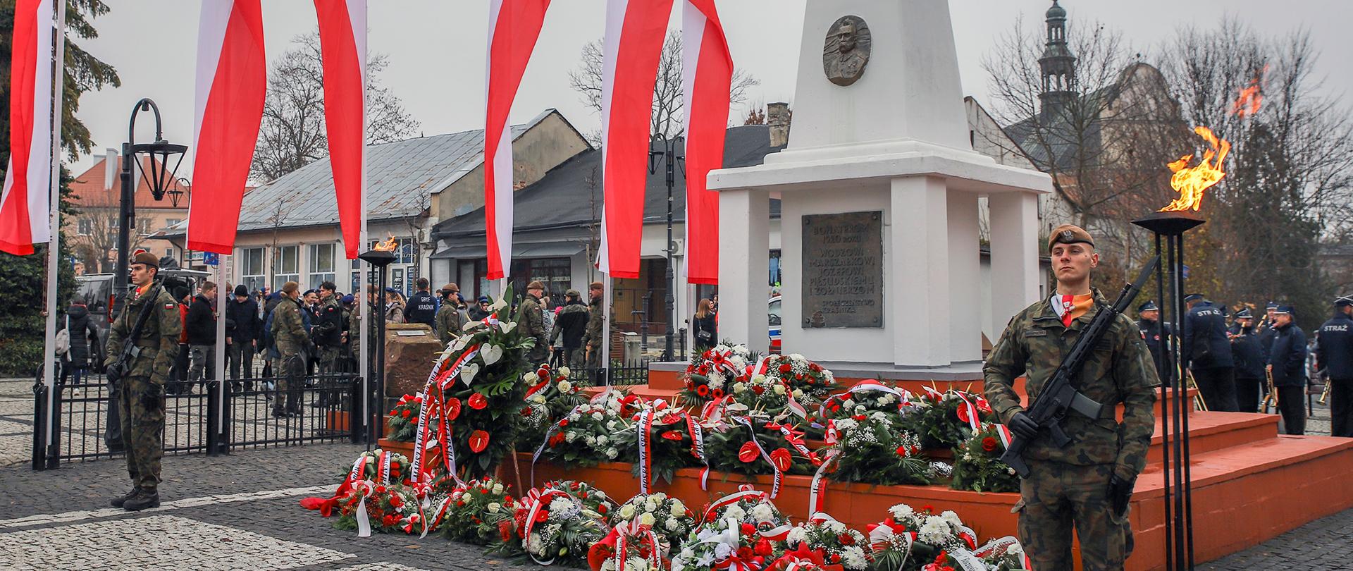 Zdjęcie przedstawia panoramę centralnego miejsca obchodów Narodowego Święta Niepodległości w Kraśniku. Na pierwszym planie stoją żołnierze w mundurach polowych pełniących wartę przy pomniku. Centralnym punktem zdjęcia jest biały, obeliskowy pomnik poświęcony Marszałkowi Józefowi Piłsudskiemu, na którego cokole widnieje tablica pamiątkowa. U stóp pomnika leży wiele wieńców i wiązanek kwiatów z biało-czerwonymi wstążkami, złożonych podczas uroczystości. W tle widać rzędy biało-czerwonych flag narodowych powiewających na masztach. Za pomnikiem i flagami rozciąga się panorama zabudowań miejskich Kraśnika, w tym wieża kościoła, oraz nagie drzewa. Scena odbywa się na brukowanym placu w pochmurny, jesienny dzień, a całość oddaje podniosły i uroczysty charakter obchodów.