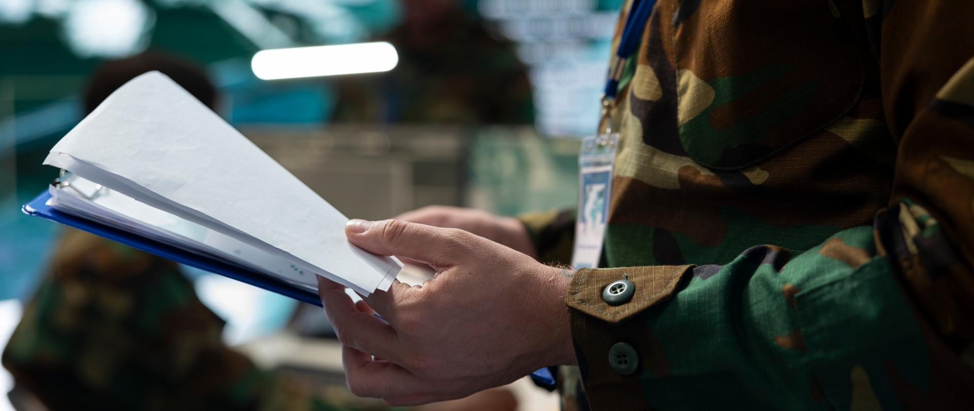Zbliżenie na osobę w mundurze w kamuflażu, trzymającą otwarty segregator z dokumentami. W tle, w rozmyciu, widoczne są inne osoby w podobnych mundurach oraz wnętrze pomieszczenia.