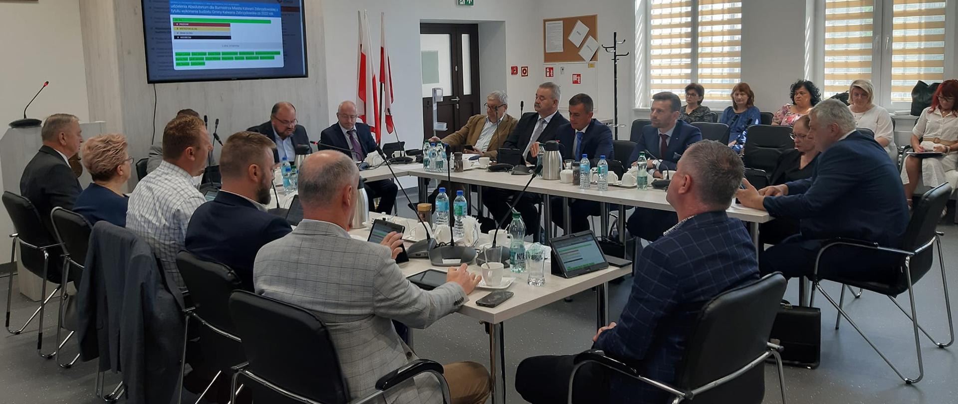 Sala sesyjna, przy stole siedzą kobiety i mężczyźni. 