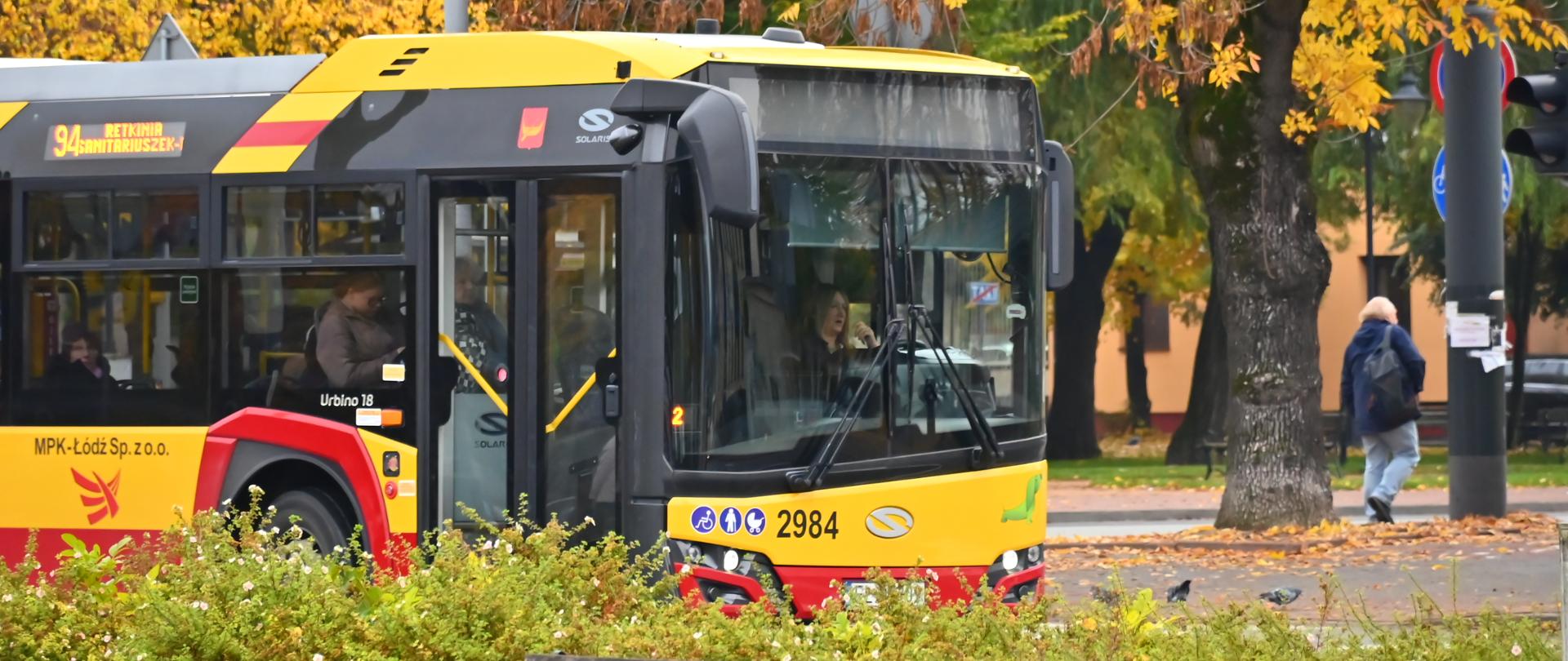 Nowoczesny autobus MPK Łódź linii 94 zatrzymujący się przy ulicy wśród jesiennych drzew; w tle pieszy przechodzący chodnikiem i sygnalizacja świetlna.