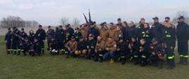 Na fotografii osoby biorące udział w zawodach strzeleckich. Część osób ubrana jest w czarne stroje z odblaskowymi paskami, druga część w brązowe kurtki i dżinsowe spodnie. Osoby siedzą na trawie. za osobami szary betonowy płot, w oddali drzewa. 