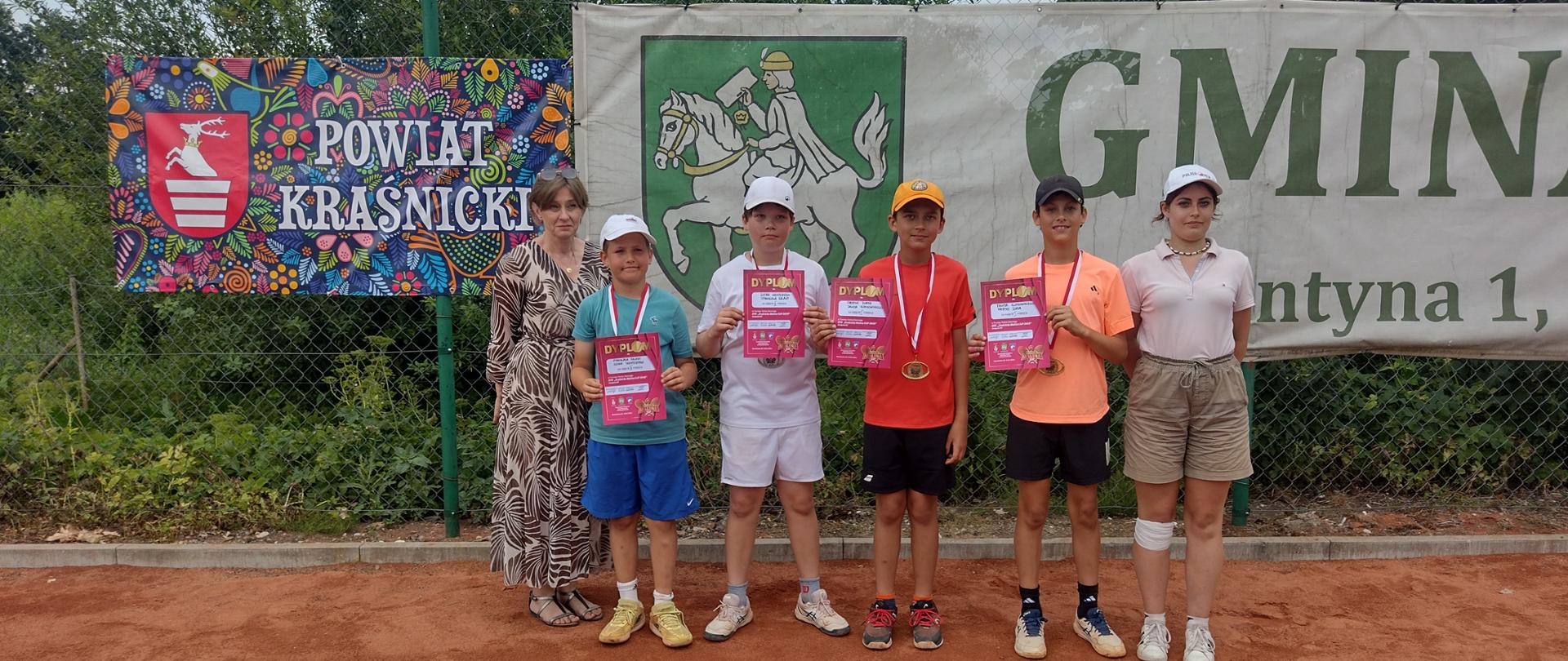 Cztery młode zawodniczki stoją w szeregu na terenie kortu tenisowego. W rękach trzymają dyplomy, a na szyjach mają zawieszone medale. Po ich lewej stronie stoi sekretarz Uczniowskiego Tenisowego Klubu Sportowego w Dzierzkowicach Aneta Piekarz.