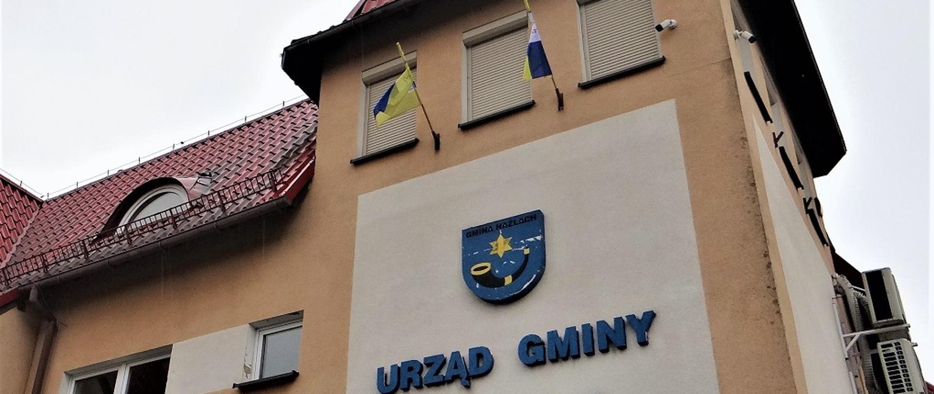 Widok na fragment frontowej ściany budynku Urzędu Gminy Hażlach. Widoczny napis wielkimi literami Urząd Gminy Hażlach. Powyżej herb gminy, nad nim, poniżej okien, dwie flagi Euroregionu Śląsk Cieszyński na drzewcach. 