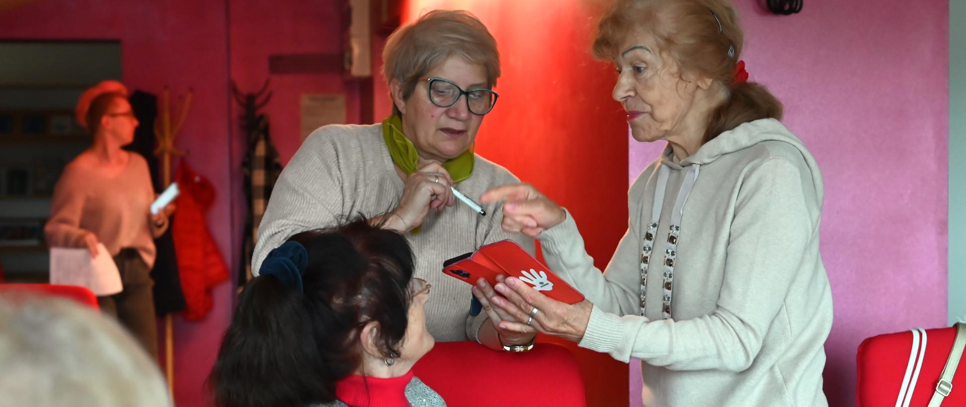 Trzy seniorki podczas zajęć. Dwie osoby trzymają telefon, pokazując na jego ekran.