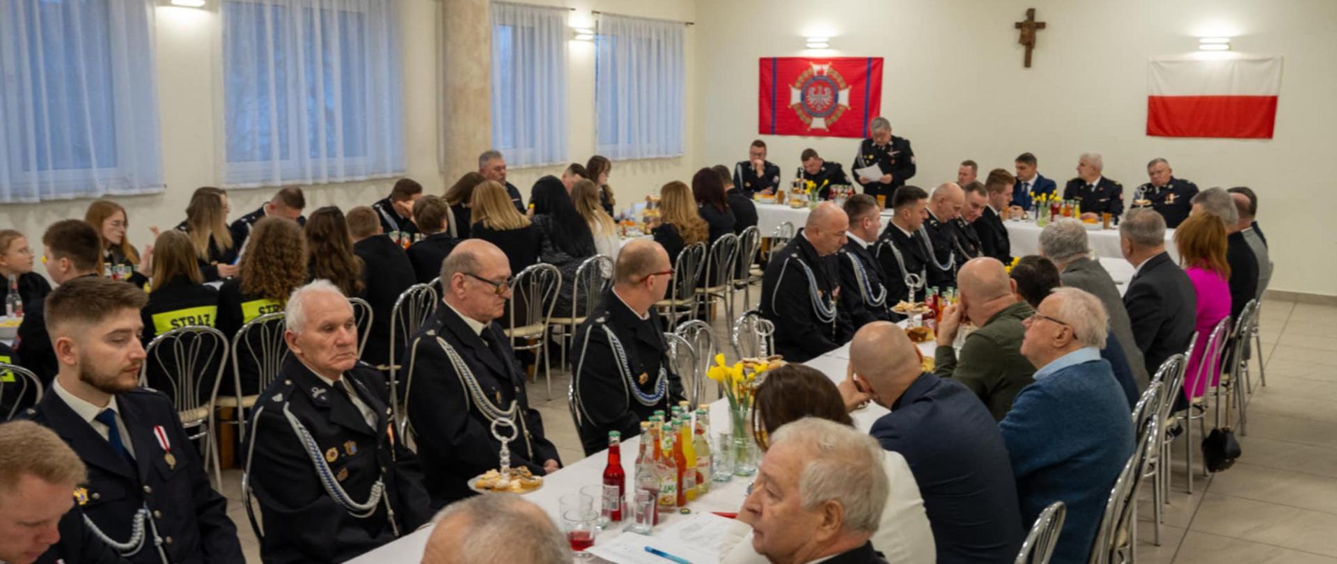 Sala spotkań wypełniona druhami Ochotniczej Straży Pożarnej oraz zaproszonymi gośćmi, shownymi przy długich stołach podczas zebrania. Strażacy ubrani w galowe mundury siedzą obok siebie, na stołach widoczne są napoje i poczęstunek. W tle wiszą sztandar OSP, krzyż oraz flaga Polski, co podkreśla uroczysty charakter wydarzenia.