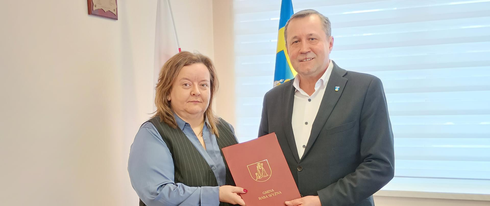 Dyrektor Małgorzata Obrochta z Wójtem Gminy Raba Wyżna w chwili przekazania umowy. Oboje patrzą w stroję aparatu w tle widać stojak na flagi oraz okno z zasłoniętą kremową roletą. 