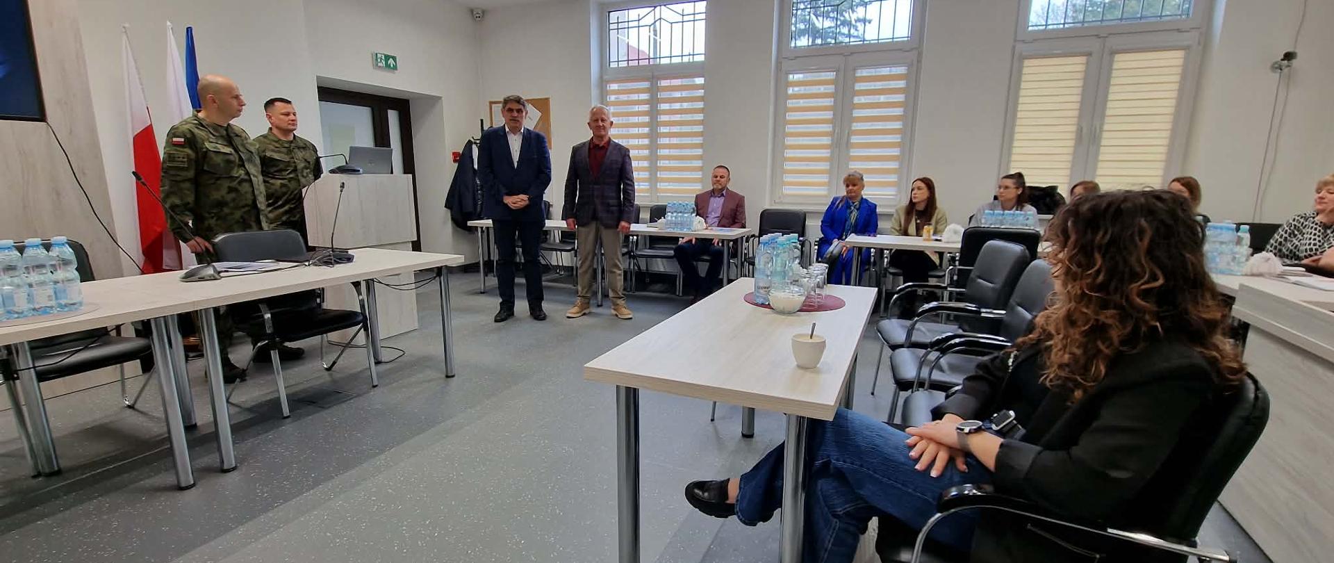Sala konferencyjna w urzędzie podczas spotkania z udziałem mieszkańców i przedstawicieli instytucji. Na pierwszym planie stoły ustawione w układzie podkowy, na nich butelki wody i filiżanki. Po lewej stronie stoją dwaj żołnierze w mundurach oraz dwóch mężczyzn w garniturach, jeden z nich przemawia. W tle widoczne okna z roletami oraz flaga Polski i Unii Europejskiej. Po prawej stronie uczestnicy siedzą na krzesłach i słuchają wystąpienia.