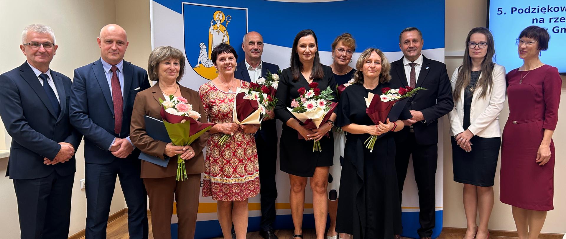 Grupa osób pozuje do zdjęcia. W tle znajduje się niebiesko-żółta dekoracja z herbem.