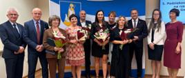 Grupa osób pozuje do zdjęcia. W tle znajduje się niebiesko-żółta dekoracja z herbem.