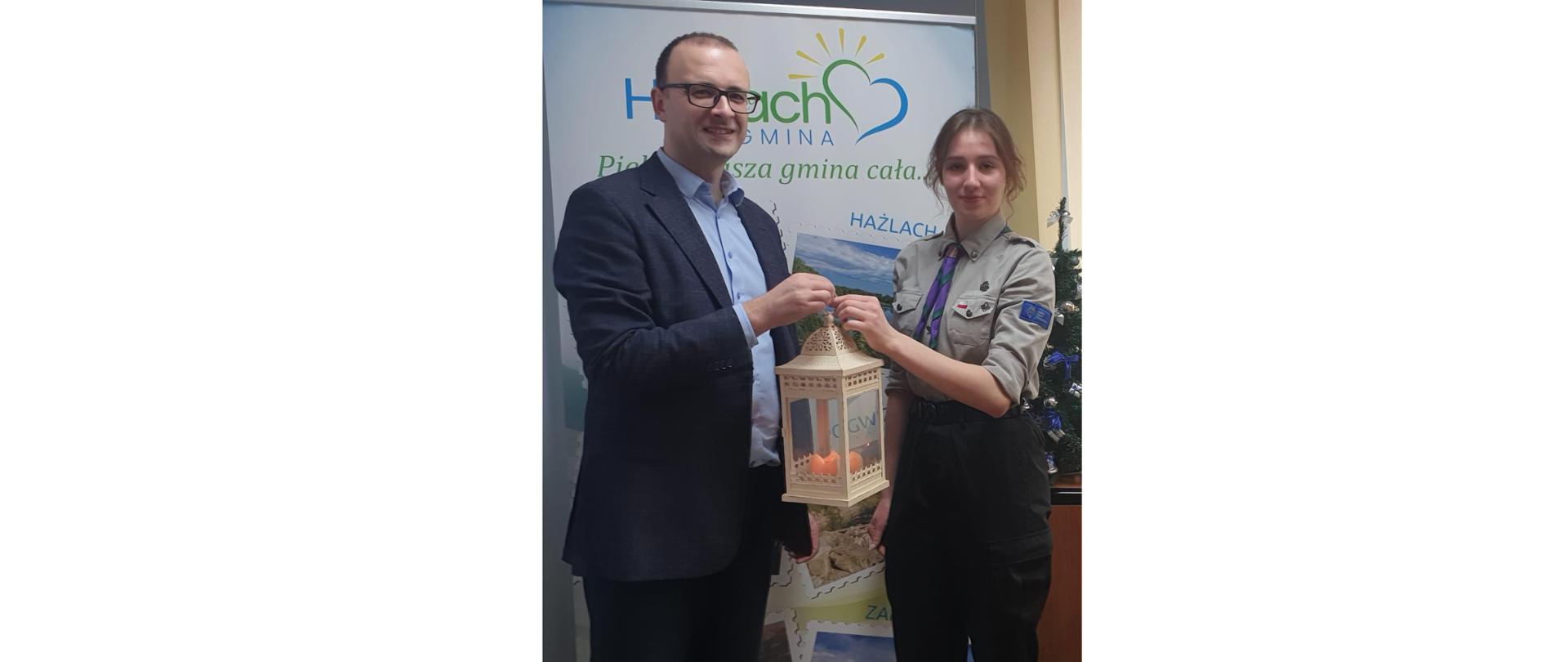 Wójt Grzegorz Sikorski przyjmuje Betlejemskie Światło Pokoju od Zofii Surzyckiej z 47 CDH „Hestia”. Dwie osoby na zdjęciu trzymają wspólnie latarnię. Za nimi baner reklamowy z logiem Gminy Hażlach 