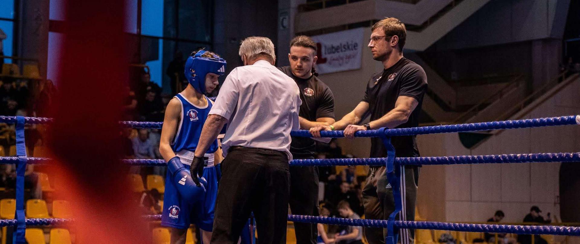 Ryszard Stefaniak w stroju bokserskim, kasku i rękawicach w kolorze niebieskim stoi na ringu naprzeciwko sędziego. Zaraz poza ringiem pierwszy od prawej stoi trener Michał Lenartowicz