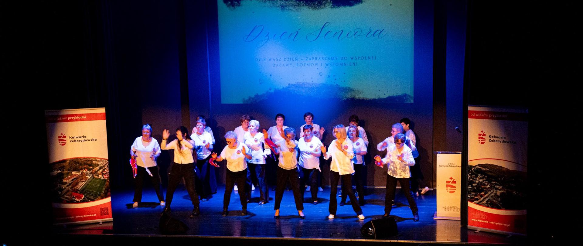 Na scenie sali widowiskowej występuje grupa seniorek ubranych w białe bluzki i czarne spodnie. Wykonują taneczny układ choreograficzny przy kolorowym oświetleniu. W tle znajduje się duży ekran z napisem „Dzień Seniora – Dziś Wasz dzień – zapraszamy do wspólnej zabawy, rozmów i wspomnień”. Po obu stronach sceny stoją roll-upy z herbem i identyfikacją wizualną Gminy Kalwaria Zebrzydowska. Widownia jest częściowo widoczna w dolnej części zdjęcia.