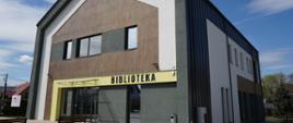 Nowoczesny budynek Gminnej Biblioteki Publicznej w Giedlarowej z widocznym napisem „BIBLIOTEKA” nad wejściem. Elewacja w odcieniach szarości i bieli, architektura nawiązująca do tradycyjnej zabudowy z nowoczesnym wykończeniem. Przed budynkiem ławki i zadbany chodnik.