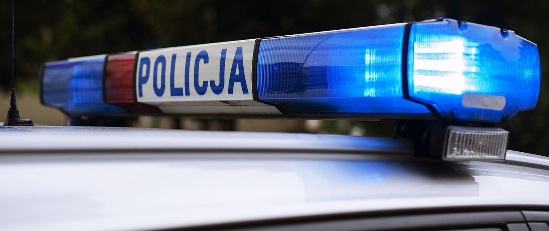 Dach samochodu na którym znajdują się migające światła policyjne w kolorze niebieskim. Na środku lamp jest umieszczony napis "POLICJA"