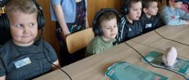 Dzieci siedzą za biurkiem, mają na uszach słuchawki