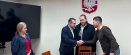 Grupa 4 osób witająca się, stojąca za brązowym stołem z krzesłami. W tle na ścianie Godło Polski, a z lewej strony telewizor.