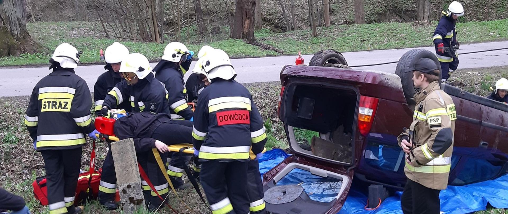 Grupa strażaków ochotników przenosząca ranną osobę z samochodu osobowego 