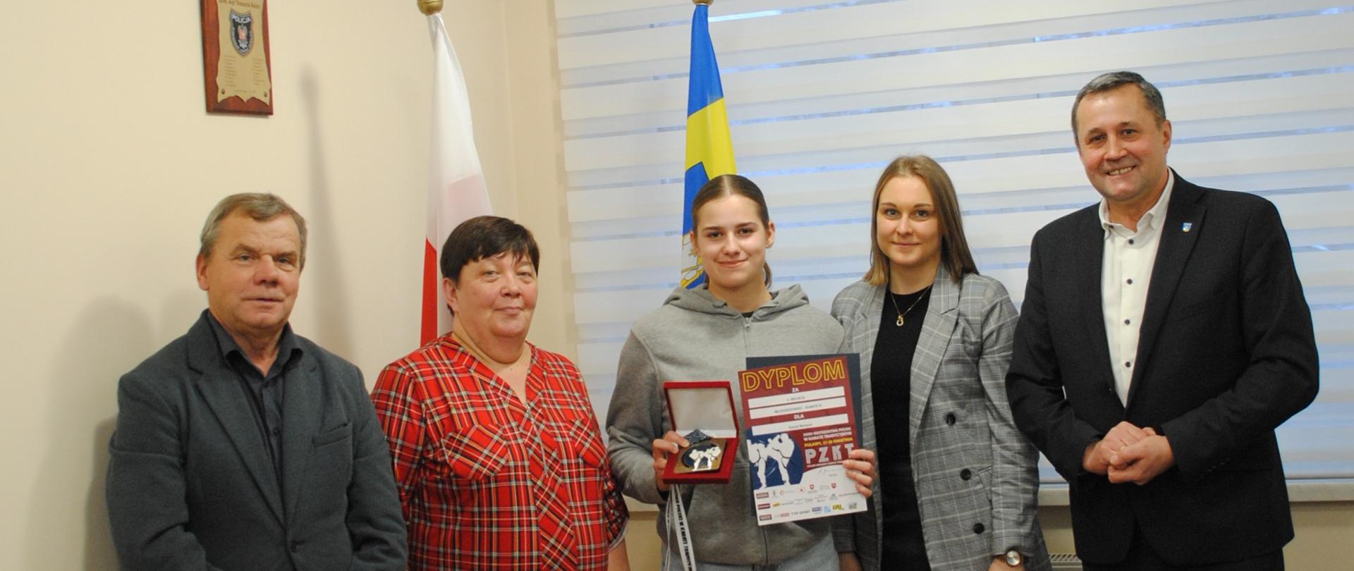 Radni i Wójt Gminy Raba Wyżna oraz zawodniczka Martyna Gacek stoją w gabinecie Wójta. Dziewczyna w rękach trzyma dyplom oraz medal. W tle widać dwie flagi