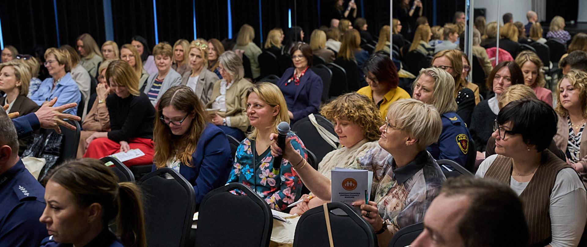 Sala wypełniona uczestnikami konferencji, część osób notuje, jedna z uczestniczek trzyma mikrofon, przygotowując się do zadania pytania.