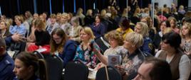 Sala wypełniona uczestnikami konferencji, część osób notuje, jedna z uczestniczek trzyma mikrofon, przygotowując się do zadania pytania.