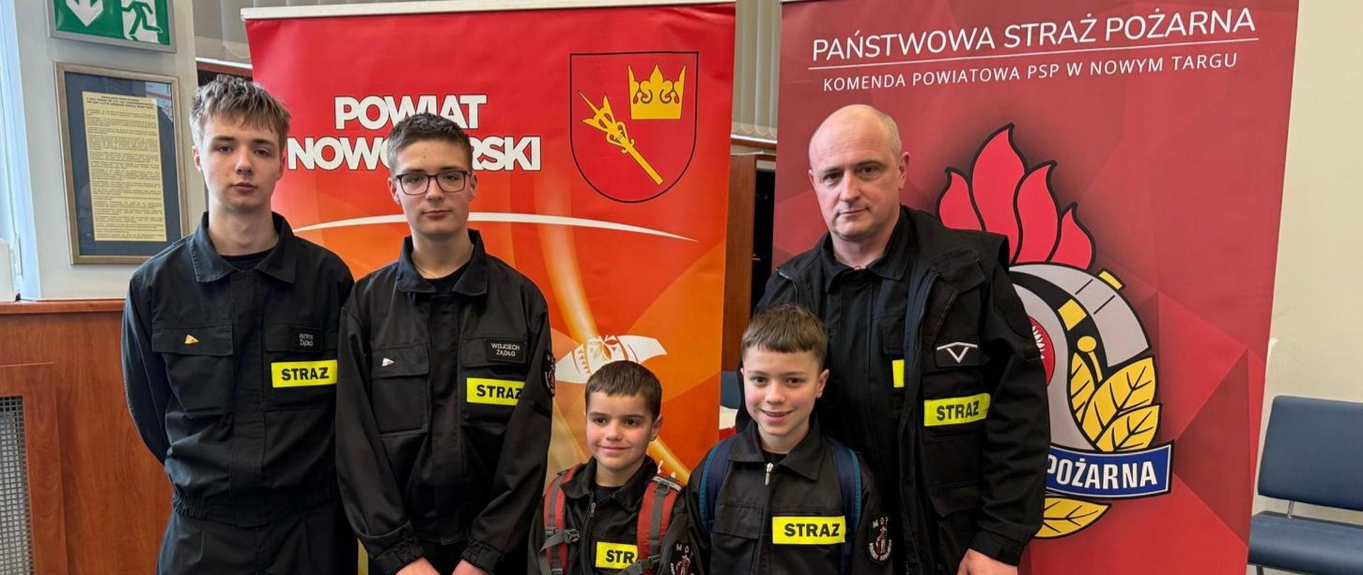 Czwórka reprezentantów z Rokicin Podhalańskich wraz z opiekunem. Stoją przed roll upami Powiatu Nowotarskiego oraz Państwowej Straży Pożarnej 