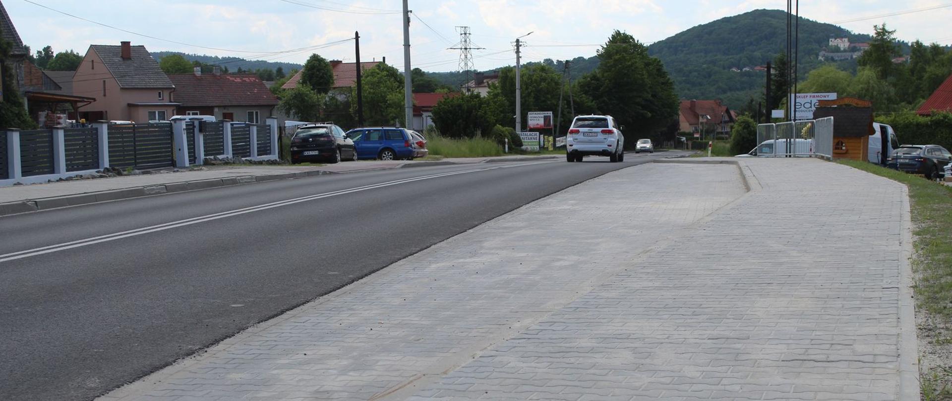 Po lewej droga krajowa nr 52 z przejeżdżającymi samochodami. Po prawej nowa zatoka autobusowa wyłożona szarą kostką brukową. W tle widać wzgórze klasztorne.