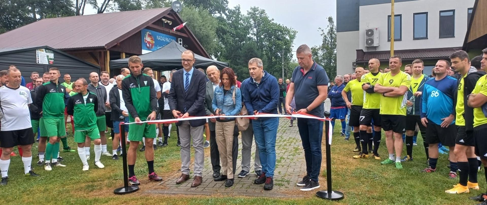 Otwarcie zmodernizowanego boiska. Biało-czerwoną wstęgę przecina pięć osób. Obok stoją zawodnicy w strojach piłkarskich. Fot. KatarzynaWijas
