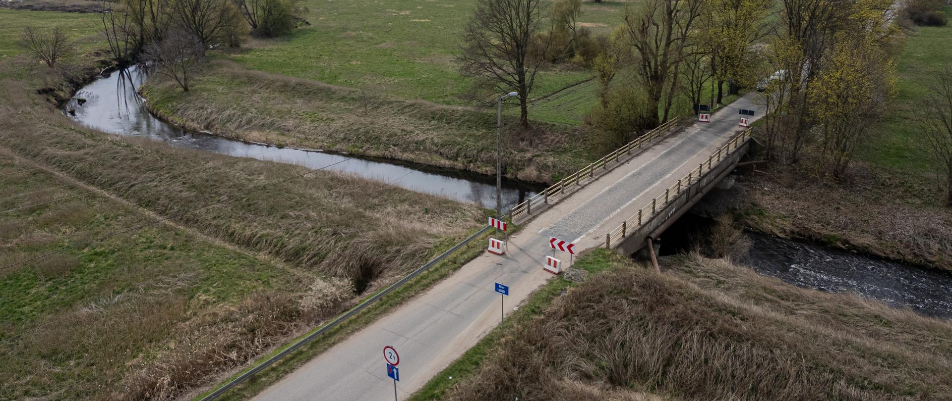Widok z lotu ptaka na wąski most w Bechcicach nad rzeką Ner, z ustawionymi ograniczeniami ruchu; w tle rozległe łąki i drzewa, a na drodze do mostu jedzie samochód.