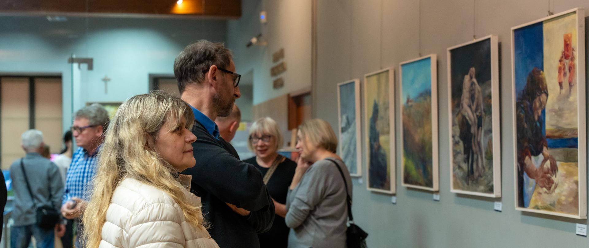Uczestnicy wernisażu oglądają obrazy zawieszone na ścianie galerii podczas wystawy malarstwa w Starym Kinie w Kalwarii Zebrzydowskiej; kilka osób stoi i rozmawia, podziwiając prezentowane prace.