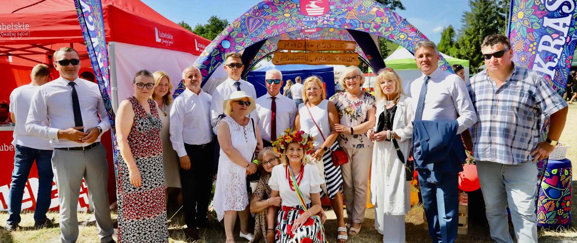 Zdjęcie przedstawia grupę ludzi pozujących do pamiątkowego zdjęcia. Przedstawiciele władz samorządowych oraz Rady Kobiet Powiatu Kraśnickiego zebrali się przed namiotem promocyjnym Powiatu Kraśnickiego